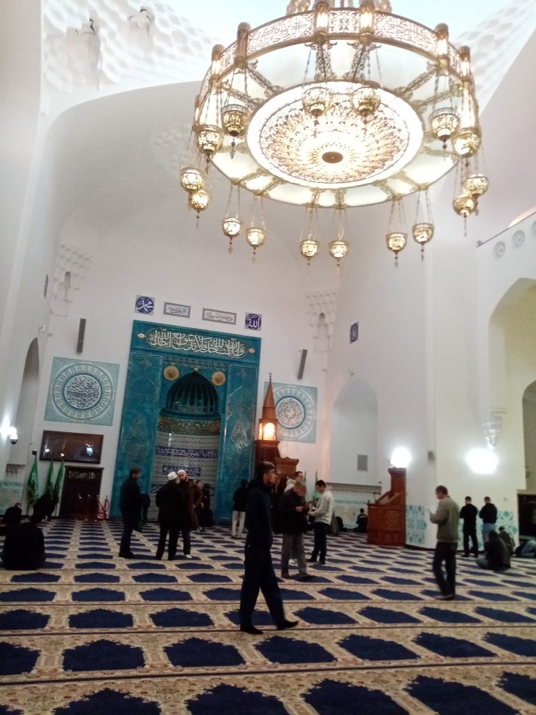 Группа экскурсантов у Соборной мечети в Санкт-Петербурге, расположенной недалеко от станции метро «Горьковская». На заднем плане — величественное здание с синим куполом и высокими минаретами в восточном стиле, украшенное керамической мозаикой и арабской вязью. Экскурсовод в национальной одежде рассказывает посетителям об истории и архитектуре мечети, соблюдая уважительную и просветительскую атмосферу