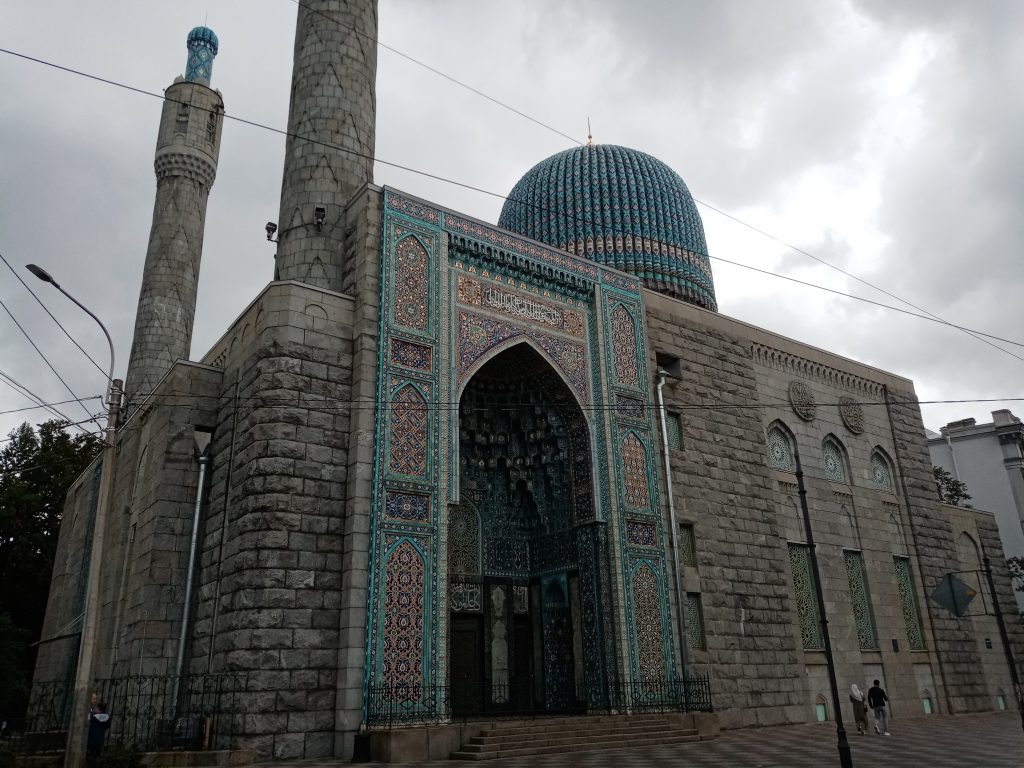 Соборная мечеть в Санкт-Петербурге Соборная мечеть в Санкт-Петербурге — величественное здание с крупным синим куполом и двумя высокими минаретами, выполненное в традиционном восточном стиле. Фасад украшен глазурованной керамикой, геометрическими узорами и арабской каллиграфией. Расположена в исторической части города, на фоне ухоженной улицы и зелени, символизируя культурное и религиозное многообразие Петербурга