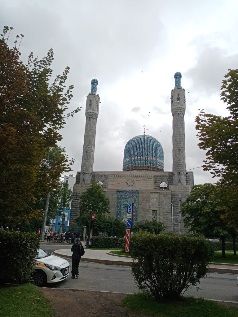 Экскурсия в мечеть Санкт-Петербурга Группа экскурсантов у входа в Соборную мечеть Санкт-Петербурга — величественного здания с синим куполом и высокими минаретами в восточном стиле. Люди в уважительной одежде внимательно слушают гида, готовясь познакомиться с историей, архитектурой и духовной жизнью одной из крупнейших мечетей Европы. На заднем плане — фасад мечети с традиционной керамической отделкой и арабской вязью